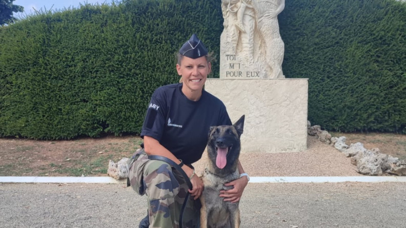 Agrandir l'image La photo représente une femme gendarme, souriante, portant un pantalon kaki, un polo et un calot noirs. La militaire est accroupie aux côtés de son chien, qu'elle tient contre elle. Derrière eux se dresse une statue blanche sur laquelle est inscrit "Toi et moi, pour eux".