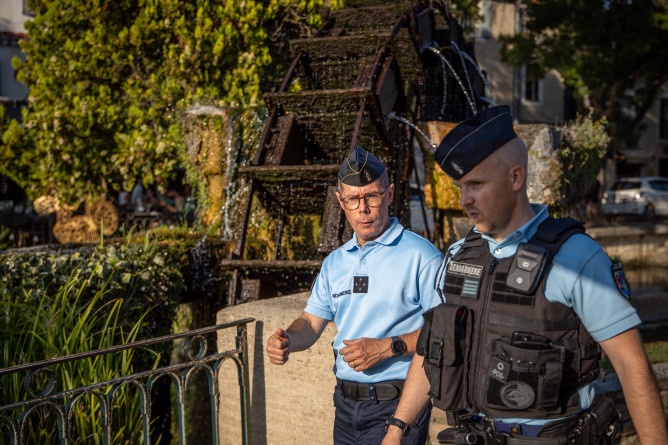 La photo représente deux gendarmes marchant côte à côte dans la rue en journée. Derrière eux, on aperçoit une roue.