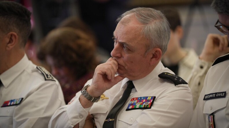 Agrandir l'image Gros plan sur le DGGN, vêtu d'une chemise blanche manches longues, cravate noire, et quatre rangées de décorations. Il est assis, entouré de deux autres personnes, également vêtues d'une chemise blanche manches longues, dont on ne voit que les épaules, et écoute attentivement, la main portée près de sa bouche.