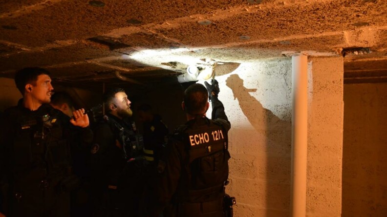 Trois gendarmes dans la pénombre d'une cave ; l'un d'eux projette la lumière de sa lampe vers une cavité dans le plafond, dans laquelle un deuxième militaire regarde.