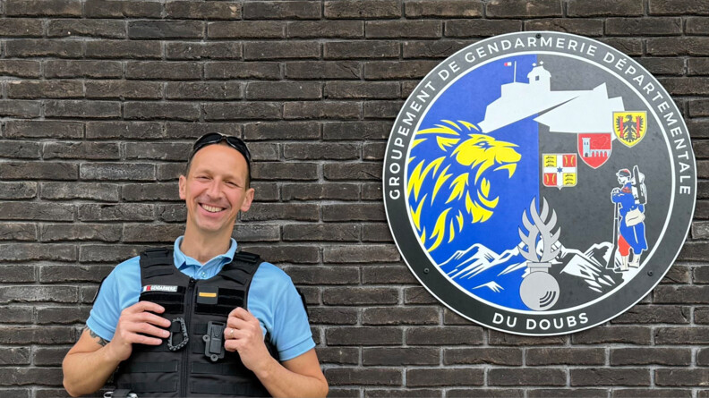 Agrandir l'image Un gendarme souriant, mes mains sur le gilet pare-balles, devant un mur en briques noires. A sa droite, accroché au mur, une rondache grandeur nature du groupement de gendarmerie départementale du Doubs