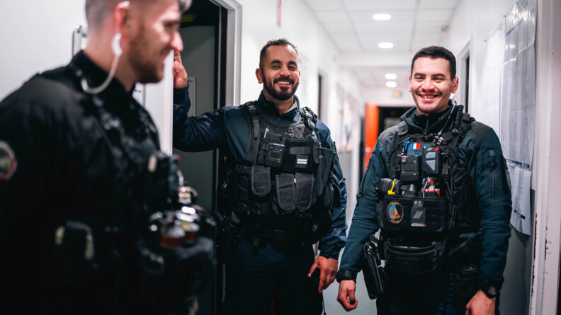 Trois gendarmes souriant en tenue d'intervention, en intérieur dans un couloir éclairé.