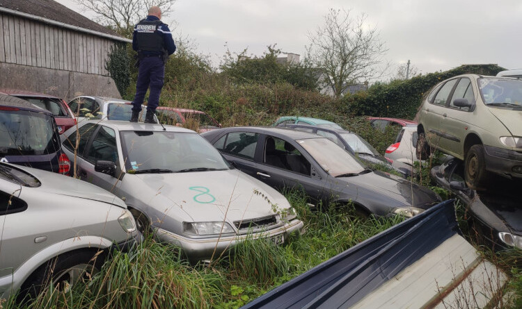 Agrandir l'image Un gendarme de dos, debout sur le toit d'une épave de voiture, au milieu d'une casse automobile dans un champ.