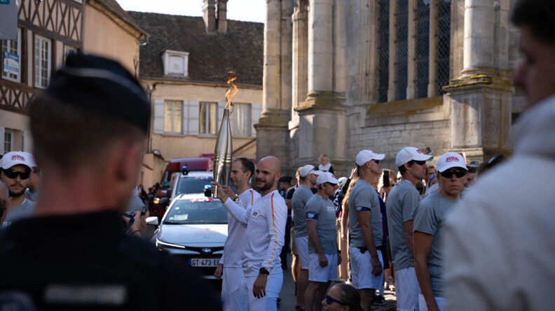 Porteur de la Flamme paralympique sous le regard d'un gendarme de dos au premier plan.
