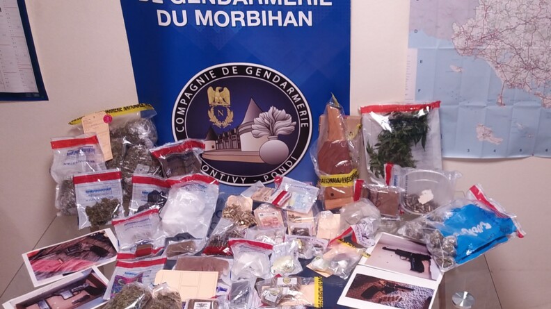Sur une table en verre sontre un mur, sous scellés, divers stupéfiants, des munitions, des photos d'armes. Entre le mur et la table, un kakemono bleu siglé du logo de la gendarmerie de Pontivy