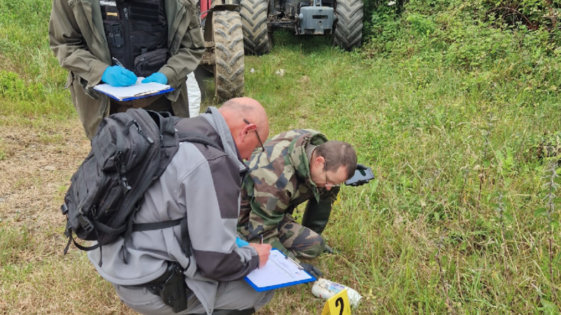 Trois hommes, penchés vers le sol, inspectent un bidon sur la pelouse. A côté de l'objet est disposé un objet un plastique avce le chiffre 2. En arrière plan, sont stationnés deux tracteurs