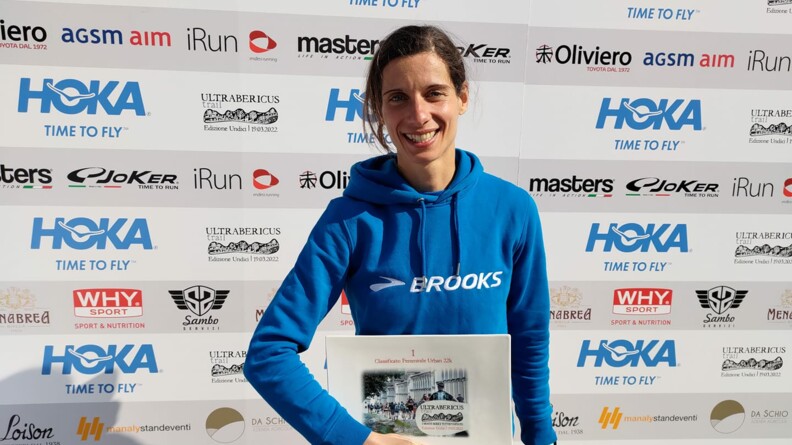 Mélina Clerc-Grosjean porte fièrement une sorte de trophée qui marque qu'elle a terminé la course première, elle est souriante et porte un pull bleu de la marque Brooks, son équipe de trail.
