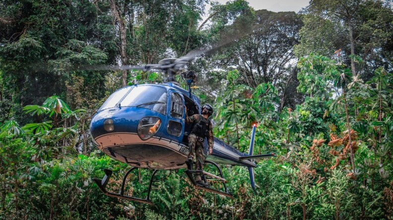 Un hélicoptère de la gendarmerie en train de se poser dans la jungle ; un gendarme en treillis avec un casque noir sur la tête sur tient debout sur l'un des patins de l'appareil.