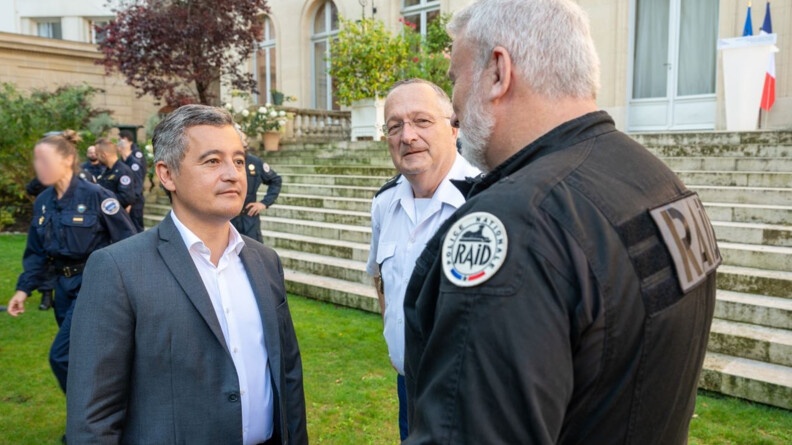 Gérald Darmanin se tient debout, dans les jardins de l'hôtel de Beauvau, devant de larges escaliers, où il discute avec un homme en uniforme noir, sur lequel est inscrite la mention RAID. A leurs côtes se trouve le directeur général de la gendarmerie nationale.
