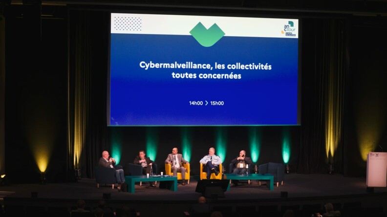 Dans un amphithéâtre plongé dans l'obscurité, excepté des lumières vertes et jaunes au fond venant du sol, une table ronde. Sur l'estrade noire, de part et d'autres, deux tables vertes. Conq hommes assis avec des micros, celui de gauche s'exprime. Deuxième en partant de la droite, le général Boget, commandant le ComCyberGend. En arrière-plan, écrit en blanc sur fond bleu roi, la thématique de la table ronde: "Cybermalveillance, les collectivités toutes concernées"