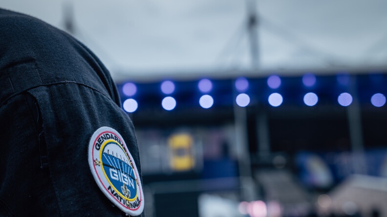 Sur l'épaule d'un gendarme en uniforme, apparait un écusson sur lequel est inscrit : Gendarmerie nationale - GIGN. On aperçoit en arrière plan les lumières floutées du Stade de France