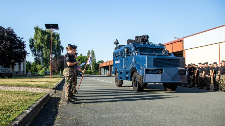 Agrandir l'image La photo représente un engin blindé de la gendarmerie nationale, de couleur bleue, avançant devant les troupes alignées au bord de la route