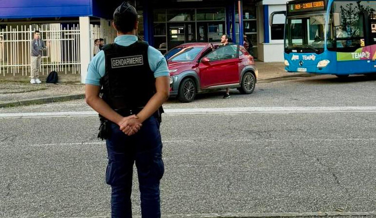 Agrandir l'image Un gendarme de dos sur la route , devant un lycée Jean Monnet, au pied duquel se trouve une voiture rouge où monte un homme et derrière elle un bus bleu