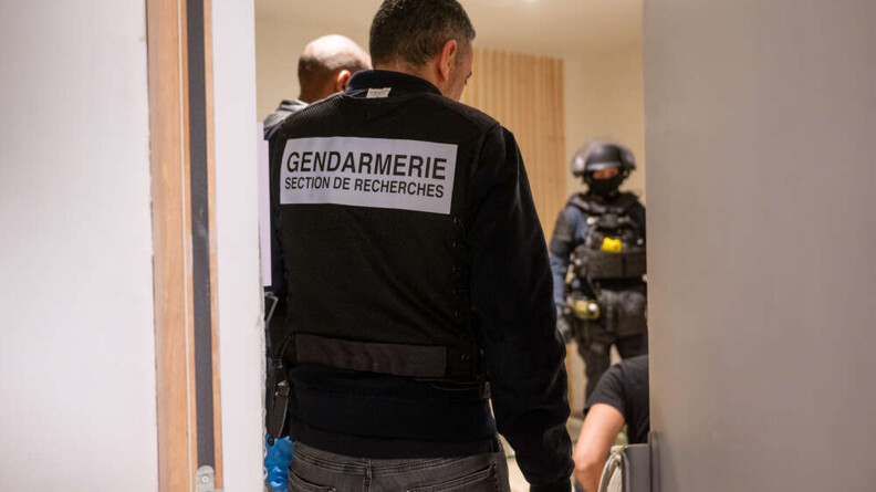 Un homme de dos, dans l'encadrement d'une porte. Il est vêtu d'un jean anthracite, d'une veste noire et d'une chasuble noire portant la mention "GENDARMERIE SECTION DE RECHERCHES" écrite en noir dans un rectangle blanc au milieu du dos. Au second plan, à gauche, on aperçoit la tête d'un homme de profil, et en bas, à droite, le bras et l'épaule d'un homme vu de dos, en T-shirt noir, que l'on imagine assis et menotté. En arrière-plan, à droite, au fond de la pièce, un gendarme mobile en tenue d'intervention, portant protections individuelles et casque.