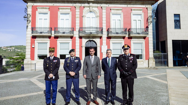 Agrandir l'image Devant un bâtiment à la façade rose, trois hommes en tenue militaire, ainsi que deux hommes en tenue civile, posent, debout, côte à côte, face à l'objectif.