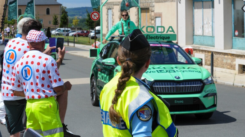 Agrandir l'image Le long de la route traversée par un véhicule de la caravane Skoda, des badauds et une gendarme de dos avec chasuble jaune