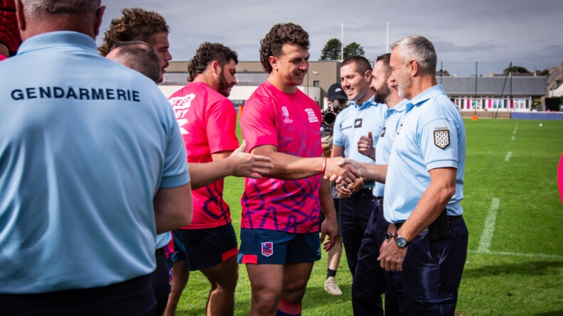 Sur la moitié gauche de l'image, 3 membres du XV du Chili, en maillot rose foncé et short bleu marine, serrent la main de trois gendarmes