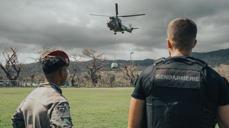 de dos un militaire au b&eacute;r&ecirc;t rouge et un gendarme en noir, un peu plus loin sur un terrain en herbe, un h&eacute;licopt&egrave;re militaire d&eacute;pose une palette de bidons d'eau. en arri&egrave;ve plan, des arbres et des collines