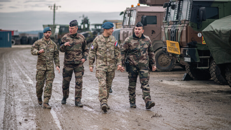 Quatre militaires alignés de gauche à droite, un gendarme néerlandais, un gendarme français, un autre néerlandais et un dernier français, dans le treillis de leur pays, echangent en marchant sur un chemin boueux, sous le ciel gris. A droite, longeant le chemin, plusieurs véhicules militaires stationnés