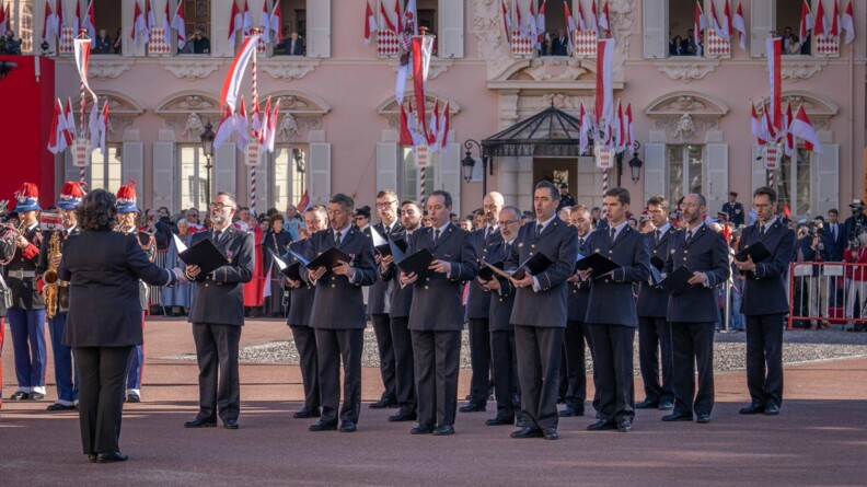 des membres du choeur de l'armée française, dirigée par leur chef, une femme, de dos, avec à gauche quelques musicien de al garde de monaco et en arriève-plan al foule e des bâtiments clairs arborant des drapeaux blancs et rouges