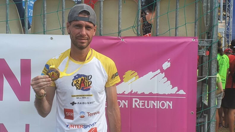 Agrandir l'image Le gendarme Maël en maillot de sport blanc portant plusieurs logos, casquette sur la tête, pose devant une bâche aux couleurs du Grand Raid de La Réunion avec sa médaille de finisher à la main.
