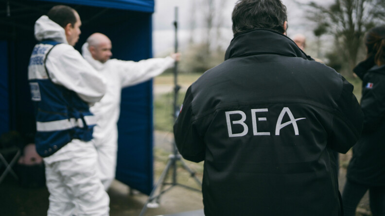 De dos, un membre du BEA, au loin floutté, deux membres des TICS dans leurs combinaisons d'intervention.