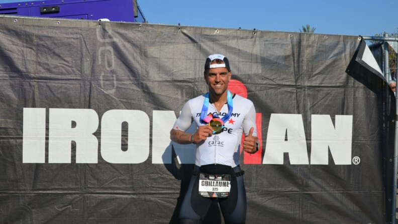 Agrandir l'image Le MDC Guillaume, revêtu d'un cycliste noir, d'un maillot blanc et casquette sur la tête, pose devant un grand panneau noir portant l'inscription IRONMAN en blanc et montre, en souriant, sa médaille de bronze.
