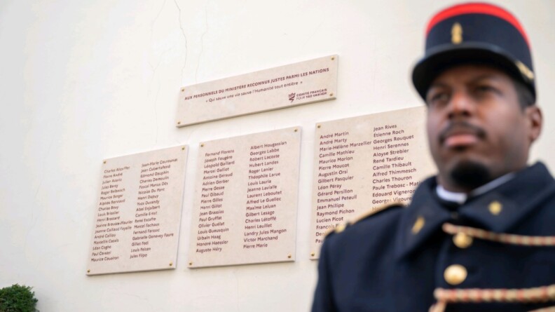 Un gendarme en premier plan en flou. Derrière lui se trouve la plaque avec tous les noms des Justes.