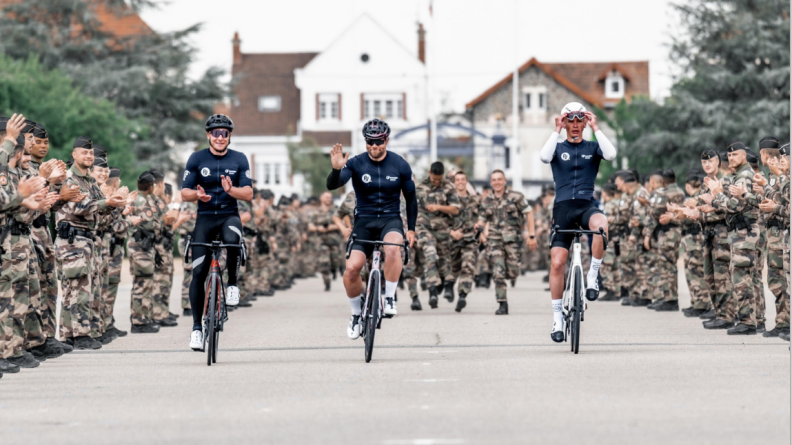 Trois cyclistes roulant sur leurs vélos vus de face en noir, celui de gauche tapant dans ses mains, celui du milieu faisant signe de la main droite, le troisième à gauche, remettant ses lunettes sur son casque. Ils sont entourés de part et d'uatres et derrière eux de très nombreux militaires en treillis