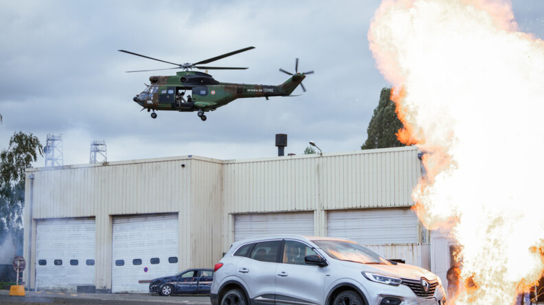 Une voiture grise passe devant une explosion qui fait une colonne de feu &agrave; droite. Dans le fond, des garages en t&ocirc;le ferm&eacute;s. Dans le ciel, un h&eacute;licopt&egrave;re Puma du GIH avec des tireurs du GIGN embarqu&eacute;s.