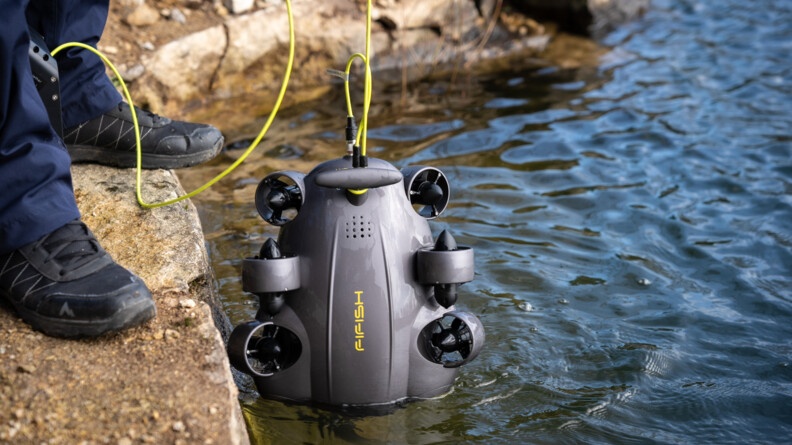 Agrandir l'image Vue sur des pieds en chaussure d'intervention au bord d'un plan d'eau, un drone subaquatique tenu par un câble jaune pénètre dans l'eau