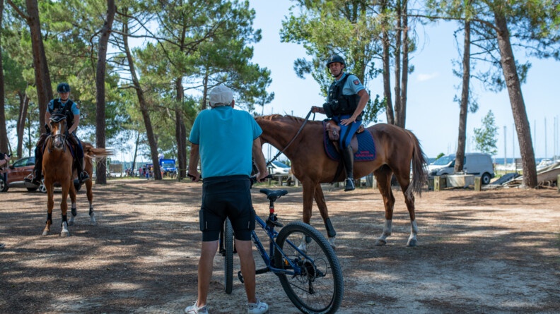 Agrandir l'image de dos au centre de l'image un homme en cycliste noir et t-shirt bleu tient son vélo et échange avec les gendarmes, montés sur leurs chevaux, dans la forêt de Biscarosse à proximité du lac. En arrière-plan on aperçoit à droite les mâts des bateaux, et la lisière du bois avec des véhicules garés