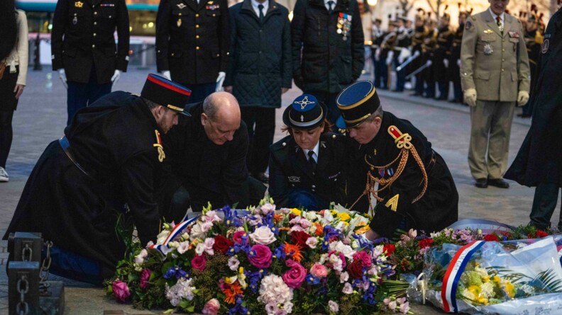 Au premier plan, un homme v&ecirc;tu de noir et trois gendarmes en grande tenue sont agenouill&eacute;s en train de d&eacute;poser une immense gerbe de fleurs sous l'Arc de Triomphe. Au second plan, cinq personnes, quatre autorit&eacute;s militaires et une civile, au garde-&agrave;-vous !