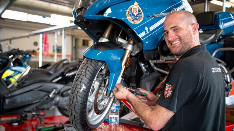 Un mécanicien répare une moto bleue de la gendarmerie. Il tient un tournevis dans la main, pointé en direction de la roue avant, et tourne la tête en souriant vers l'objectif