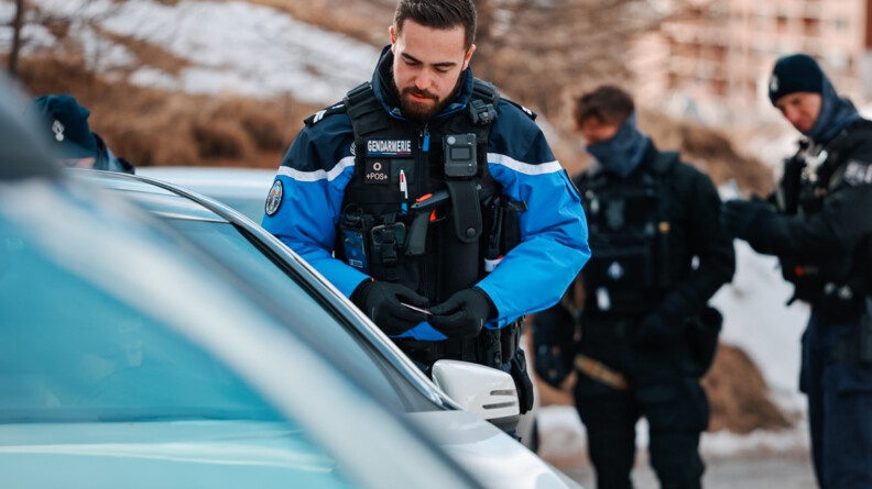 Un gendarme effectue un contrôle routier en bord de route de montagne, vue sur l'habitacle du véhicule; en arrière plan deux autres gendarmes
