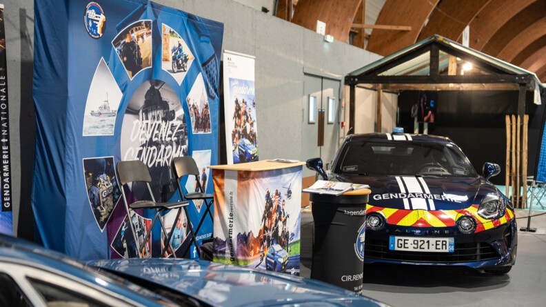 Une Alpine de gendarmerie récente exposée auprès d'un stand de recrutement de la gendarmerie nationale, avec les mentions "devenez gendarme" et "la gendarmerie recrute" et de nombreuses photos