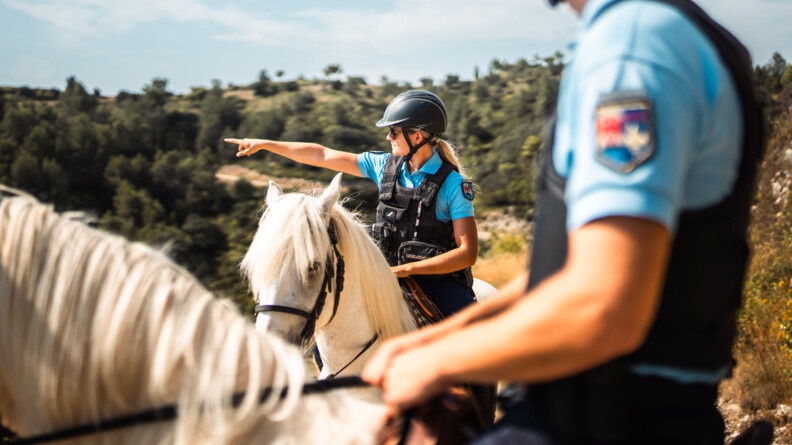 Deux gendarmes (un homme et une femme), assis sur des chevaux blancs, dans un environnement naturel, sont arrêtés. La femme désigne un point du paysage avec l'index de sa main droite.