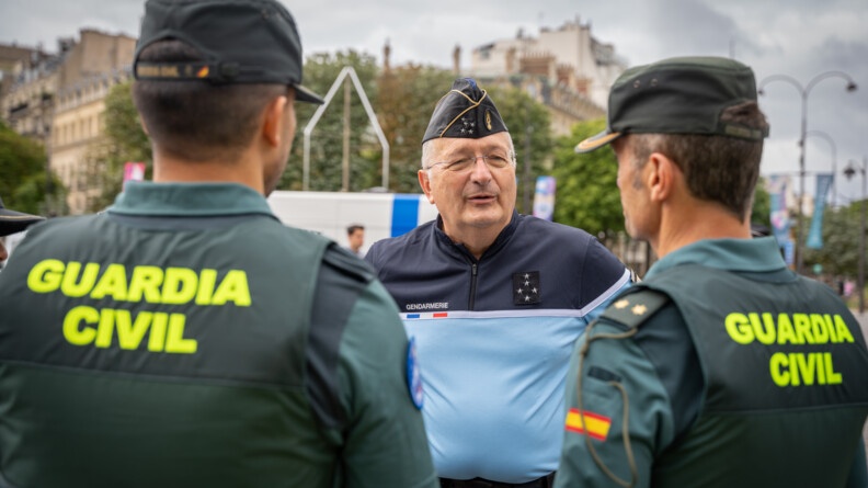 Le directeur général de la gendarmerie nationale de face, vêtu d'un polo bleu clair avec le tiers supérieur bleu nuit, calot sur la tête. Face à lui, de dos, deux gardes civils espagnols, en tenue verte, portant la mention GUARDIA CIVIL en jaune fluo sur le dos, et portant une casquette.