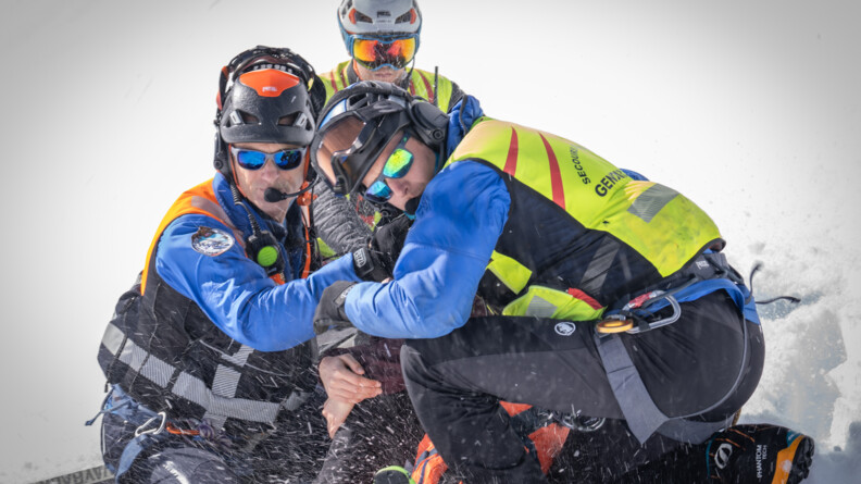 Trois gendarmes secouristes du PGHM protègent une victime de l'envolée de la neige dû à l'arrivée de l'hélicoptère.
