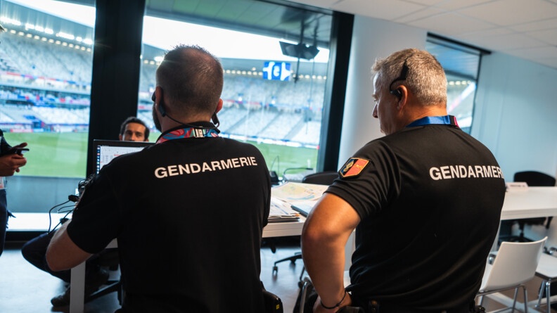 A droite, le colonel Sorre, commandant le GTG, et &agrave; gauche, un autre gendarme, &eacute;galement vu de dos. En arri&egrave;re plan, l'int&eacute;rieur du stade vu &agrave; travers une paroi vitr&eacute;e.