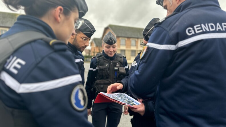Un groupe de cinq gendarmes - deux femmes et trois hommes - en tenue, se tiennent debout dans la rue, en plein jour. L'un d'eux s'exprime, un cahier &agrave; la main. On aper&ccedil;oit en arri&egrave;re plan des arbres et la fa&ccedil;ade d'un b&acirc;timent en brique rouge.