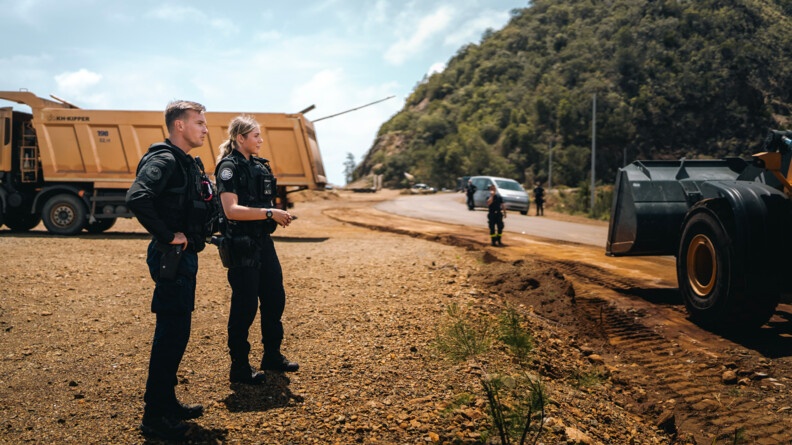 Deux gendarmes mobiles, un homme à gauche et une jeune femme à droite, vus de profil, sont au bord de la Route territoriale 3 et observent une pelleteuse à l'ouvrage. Au second plan, à gauche, un gros camion-benne jaune ; à droite, d'autres personnels sur la route.