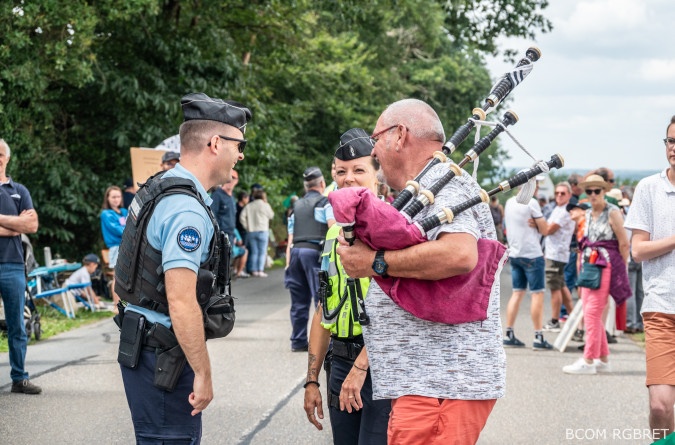 Agrandir l'image Deux gendarmes en uniforme échangent avec le sourire avec un spectateur tenant une cornemuse en tissu rose. L’ambiance est conviviale, avec plusieurs personnes en arrière-plan le long de la route, dans un décor champêtre.