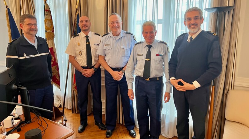 AUtour d'un genéral de gendarmerie en chemisette blanche et pantalon droit bleu marine, quatre aumôniers de aprt et d'autre devant un mur ave deux fenêtres hautes, des rideaux claors et un drapeau français