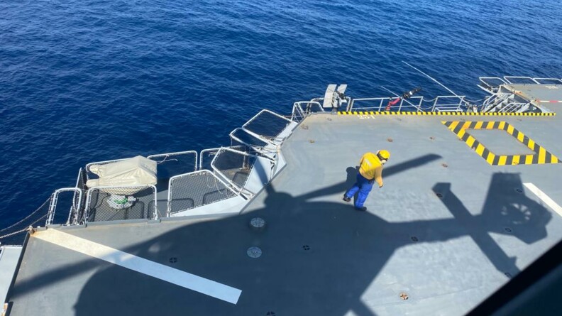 Vue sur la mer et sur le pont du porte hélicoptères Mistral, où se projette l'ombre de l'hélicoptère de la gendarmerie nationale. Sur le pont, un agent de piste vêtu d'une chasuble et d'un casque jaunes, vu de dos.