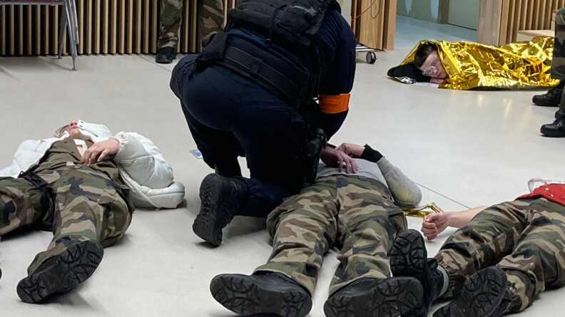 Quatre personnes incarnant des blessés sont couchées sur le sol, trois au premier plan et une au fond à droite de l'image. Un militaire du Psig Sabre, vu de dos, est accroupi à côté de celui du milieu.