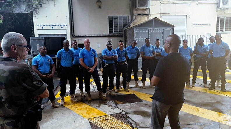 Agrandir l'image Deux hommes de dos, l'un en treillis kaki et l'autre en civil, s'adressent à une rangée de policiers seychellois vêtus de pantalons de treillis noirs et de polos bleus.
