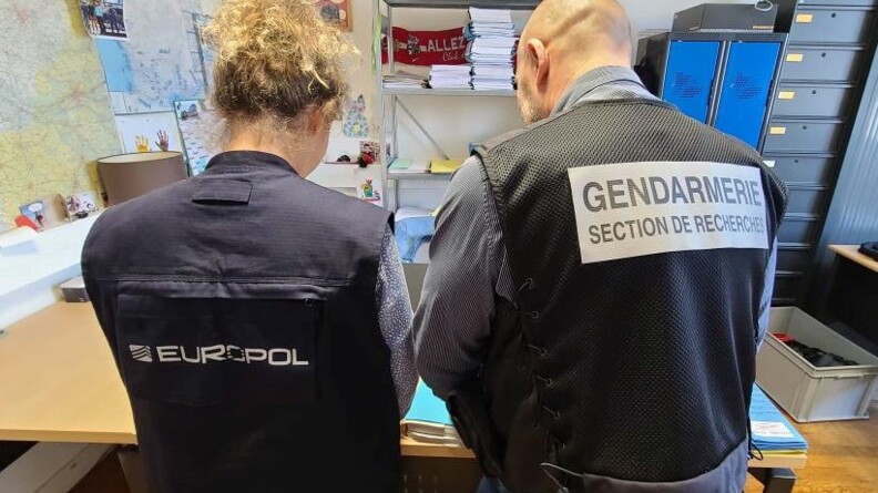 Agrandir l'image Deux personnes vues de dos, dans un bureau : à gauche, une enquêtrice portant une chasuble Europol, et à droite, un enquêteur portant une chasuble Gendarmerie Section de recherches.