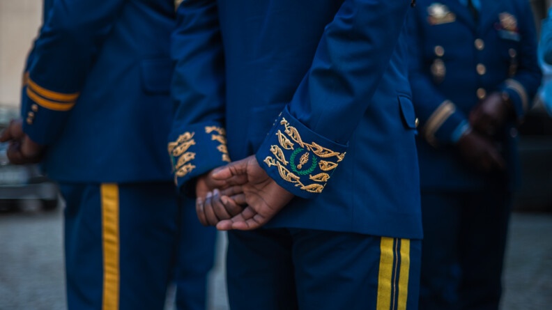 Agrandir l'image On distingue les jambres et le dos de deux gendarmes congolais de os, se tenant debout, les mains serrées dans le dos. Un gros plan est fait sur les mains de l'un deux, permettant de distinguer les broderies au bas des manches de sa veste. On aperçoit aussi, en second plan, le torse et les jambes floutés d'un troisième gendarme congolais.