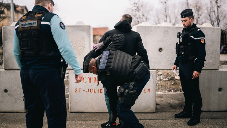 Agrandir l'image Un gendarme, entouré de deux autres gendarmes, réalise une fouille sur un homme vêtu d'un jean et d'un blouson noir, se tenant debout, de dos. La scène se déroule sur un trottoir, par temps gris.